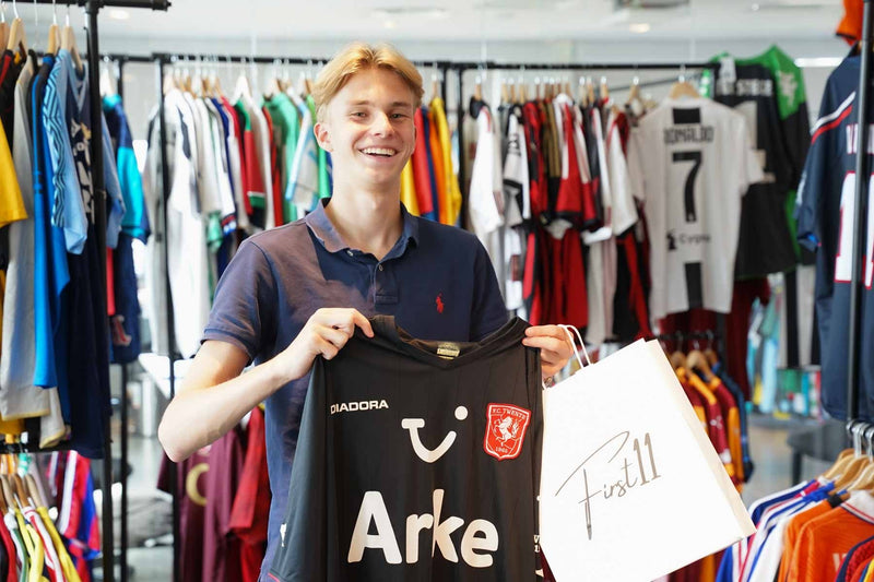 First11 founder Stefan Haas holding an FC Twente shirt and a First11 bag in front of racks of vintage football shirts in the store